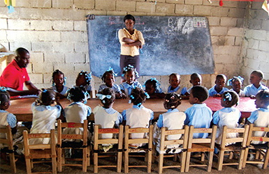 Students sit around a table and listen while a teacher stands in front of a blackboard.  